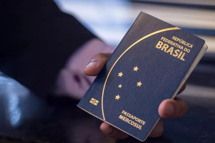 Foto de uma mão branca segurando um passaporte brasileiro. O passaporte é uma caderneta pequena de capa azul rígida, com o nome República Federativa do Brasil, um semi círculo e cinco estrelas